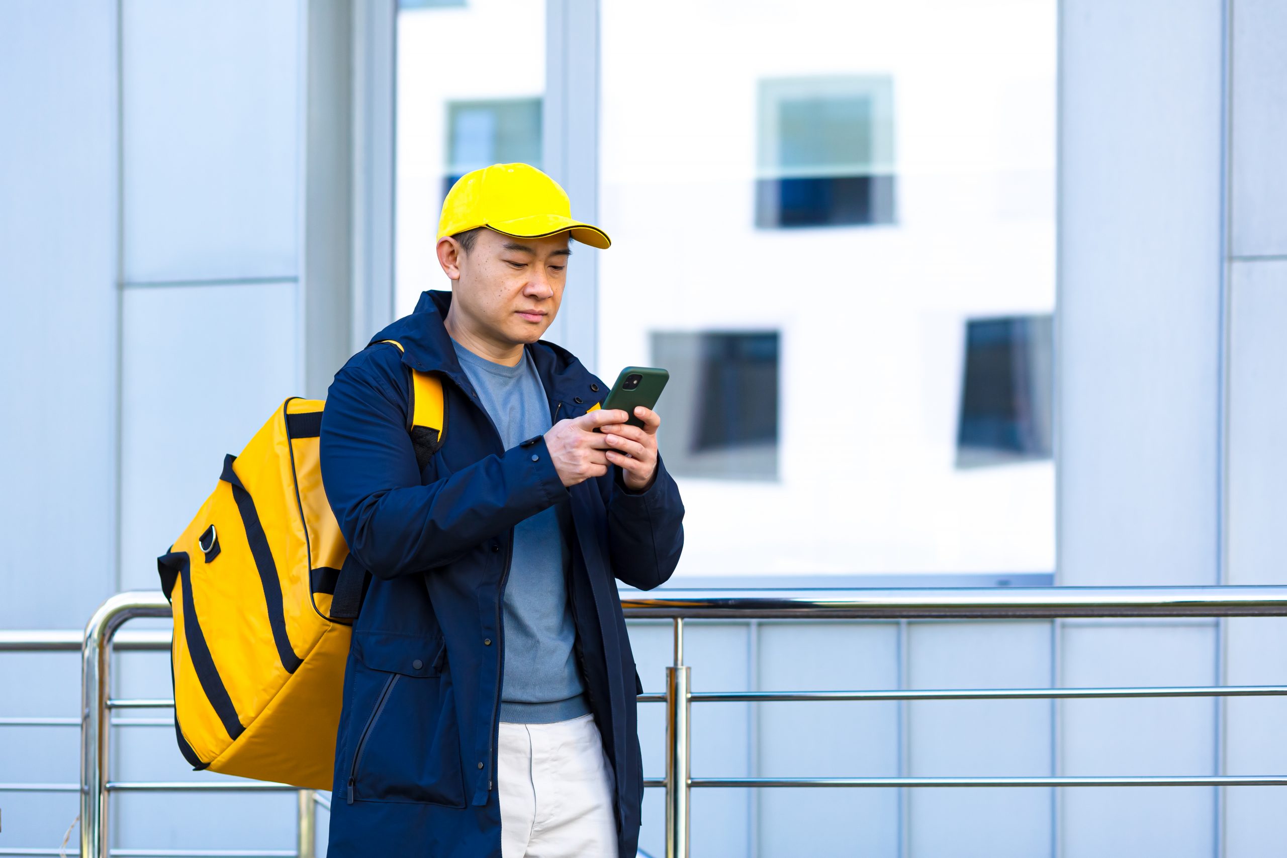 asian-male-food-courier-using-mobile-using-mobile-phone-app-smartphone-while-standing-city-street-with-insulated-with-thermal-backpack-delivering-food-looking-customer-address-purchase-online-map